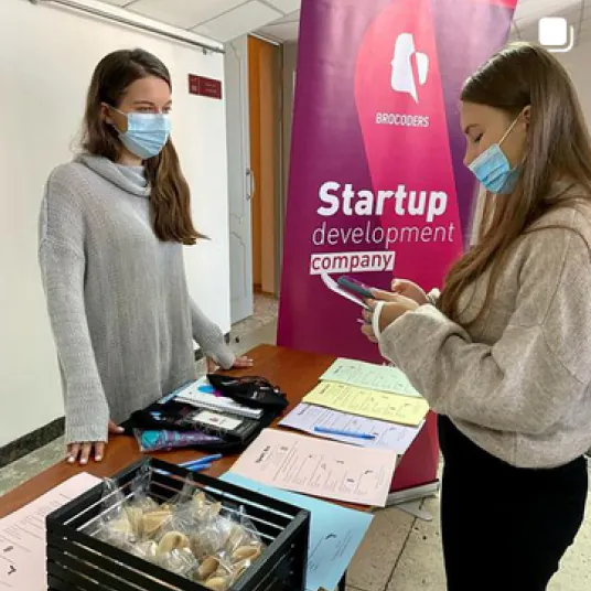 Two young women wearing face masks stand at a table with papers